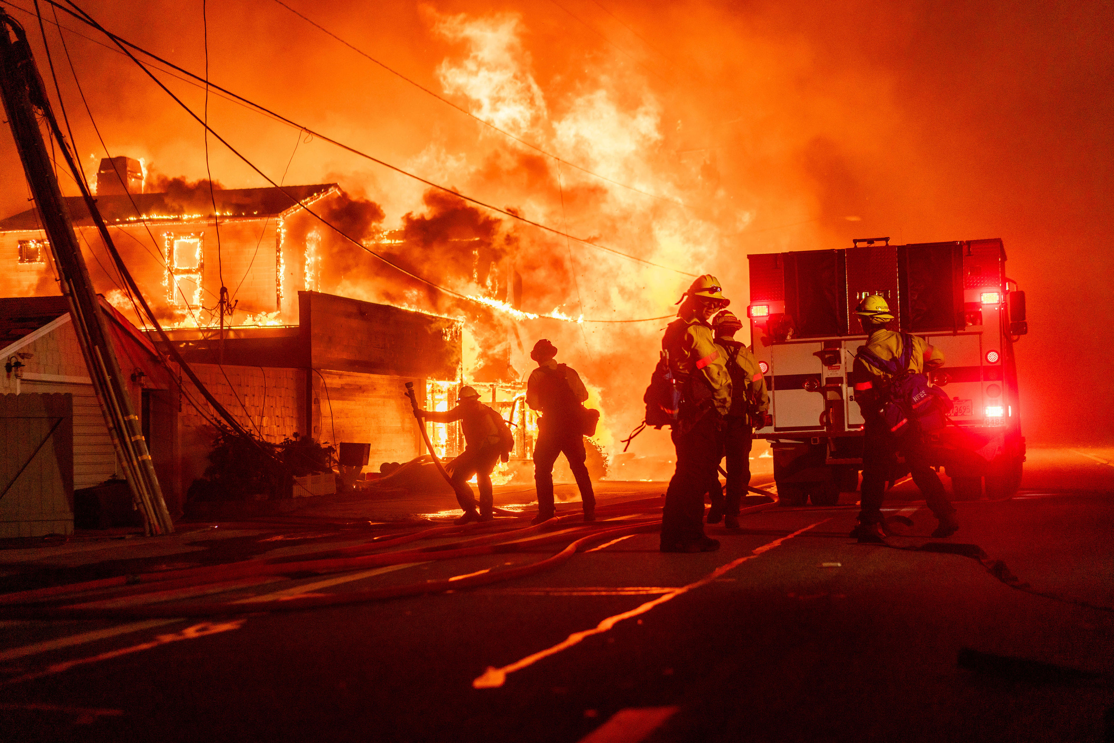 Firefighters fight flames from a house on fire with a firetruck in view