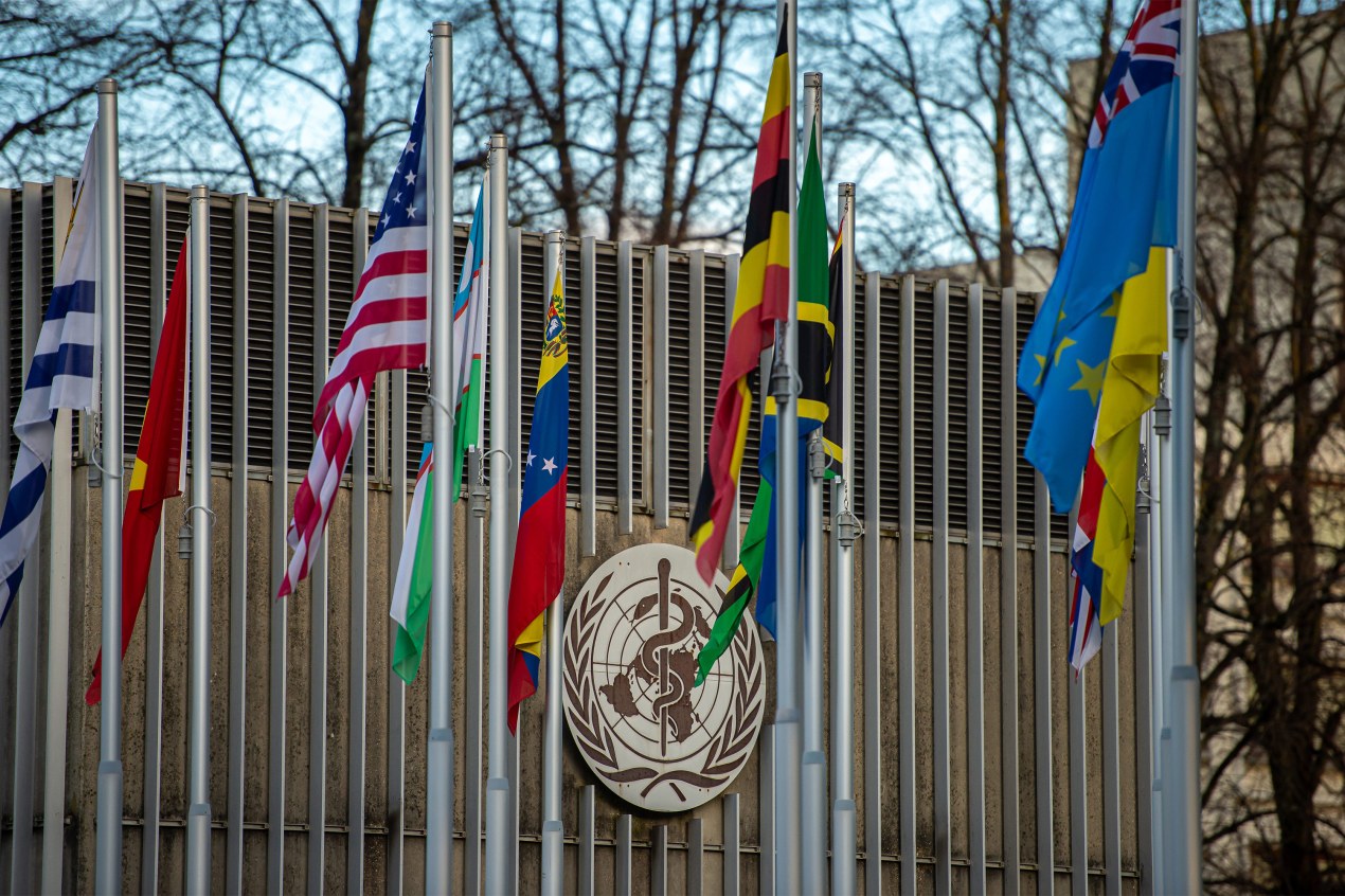 A photo of the exterior of the World Health Organization headquarters. A row a flags fills the frame in front of the building, including the American flag.