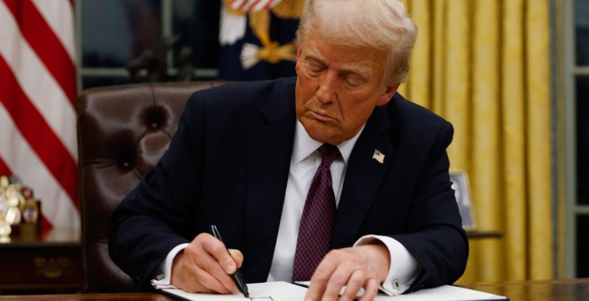 President Donald Trump signs behind a desk in the Oval Office and signs a document.