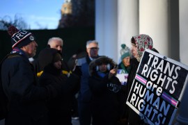 A small crowd of people face each other outdoors on a cold day. A man on the left wears a winter hat that says "TRUMP." A woman on the right holds a sign that says, "Trans rights are human rights!"