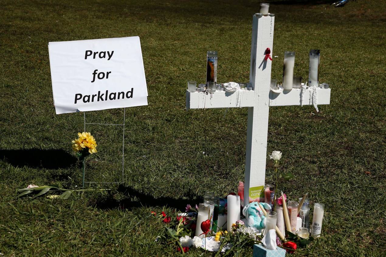 A white cross stands in green grass, adorned with candles and other memorial objects. Beside the cross is a sign that reads, "Pray for Parkland."