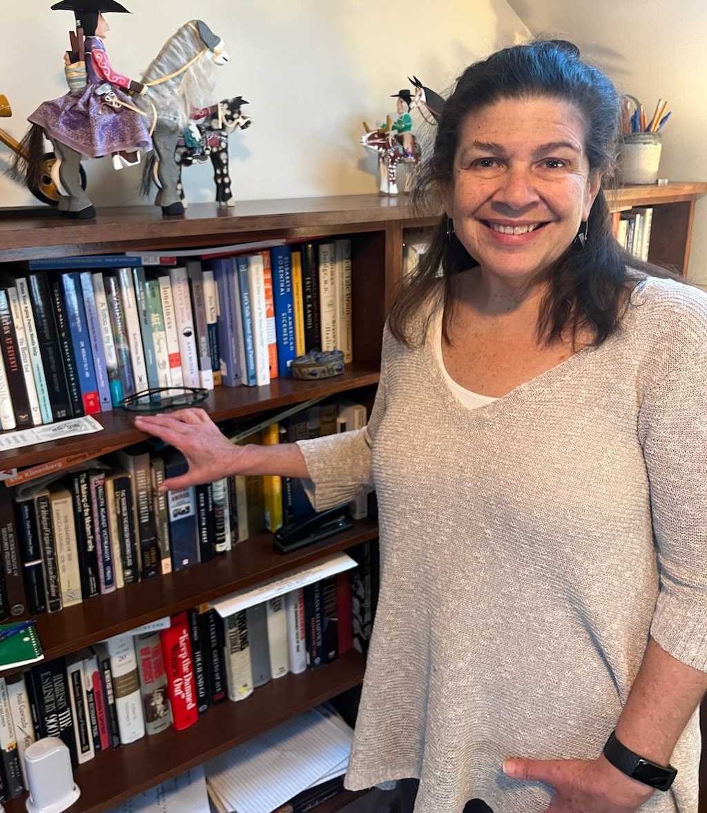 A senior woman in a grey sweater stands by a book shelf. She has shoulder length brown hair and is smiling.