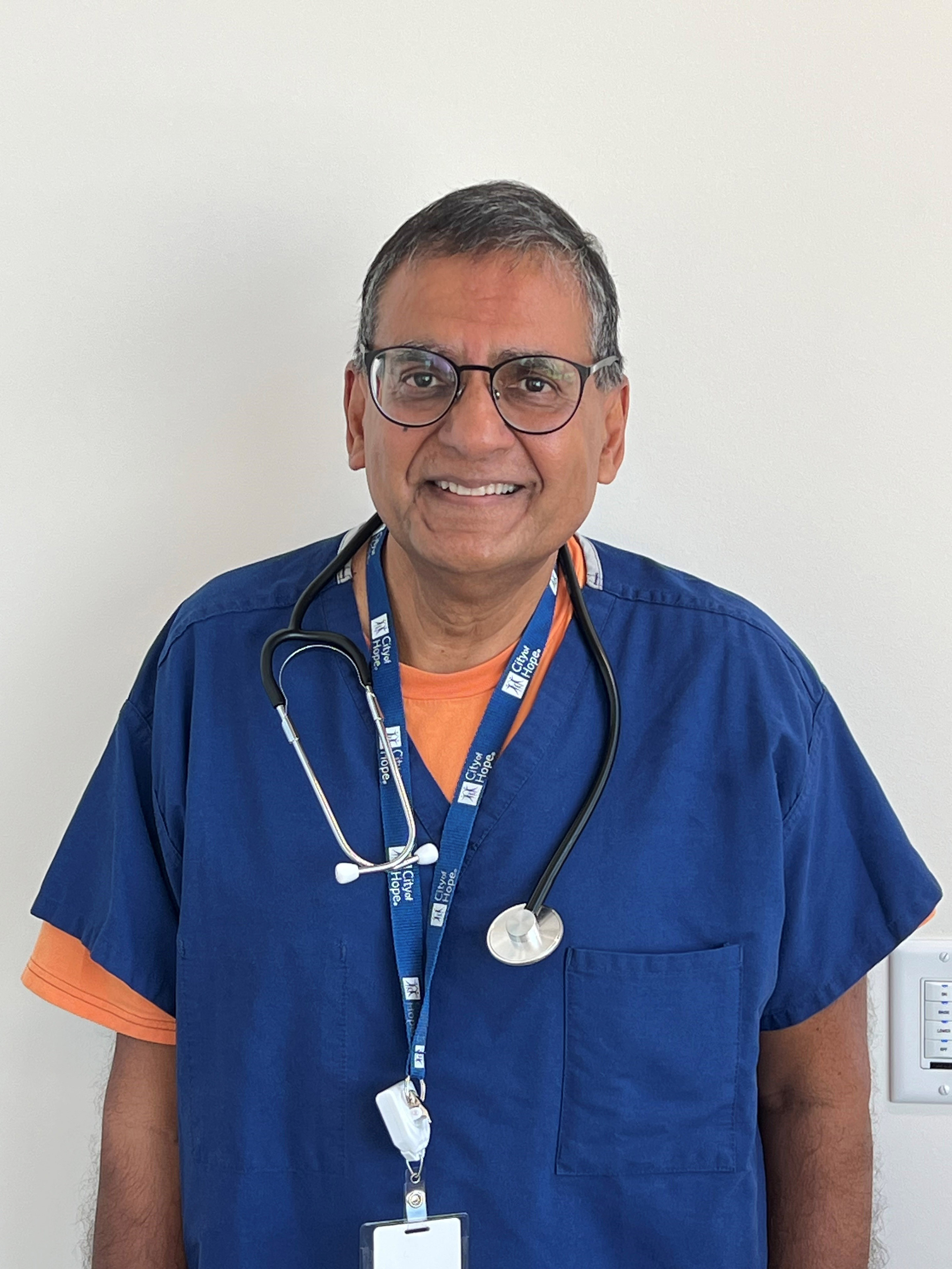 A portrait of Dr. Ravi Salgia. He wears blue scrubs and has a stethoscope hanging around his neck. He smiles at the camera.