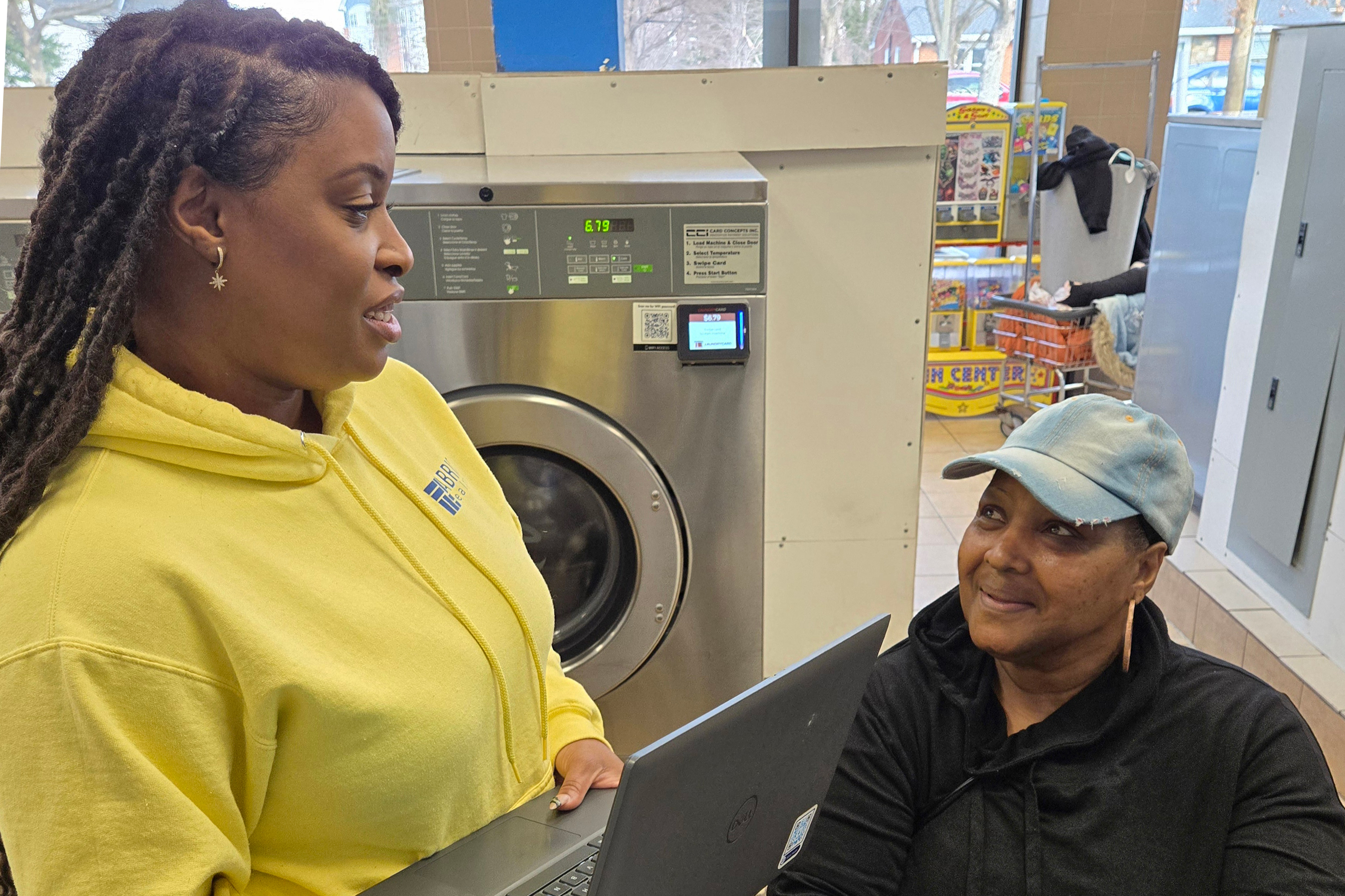 A photo of Adrienne Jones standing while holding a laptop and speaking to Patti Hayes, seated. A laundry machine is behind them.