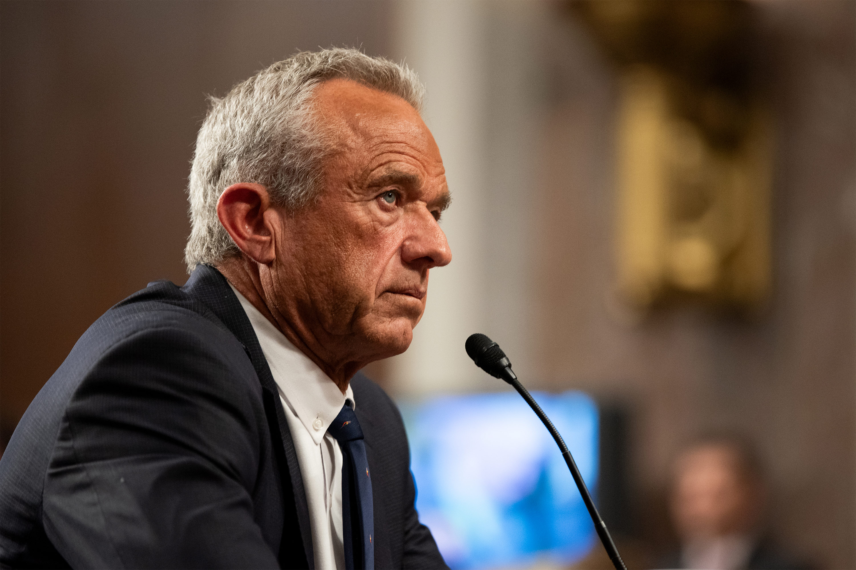 A photo of Robert F. Kennedy Jr. seated in a Senate hearing room, looking forward. A microphone is in front of him.