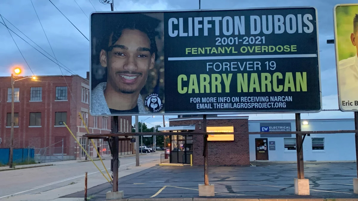 A billboard with an image of a smiling young man reads "Cliffton Dubois, 2001-2021, fentanyl overdose, forever 19, Carry Narcan, For more info on receiving Narcan email themilagrosproject.org."