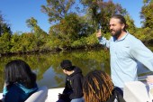 A small group of students are sitting on a small boat. An adult man stands behind the group, speaking. In the background is a river with deep green water. It is a sunny day.