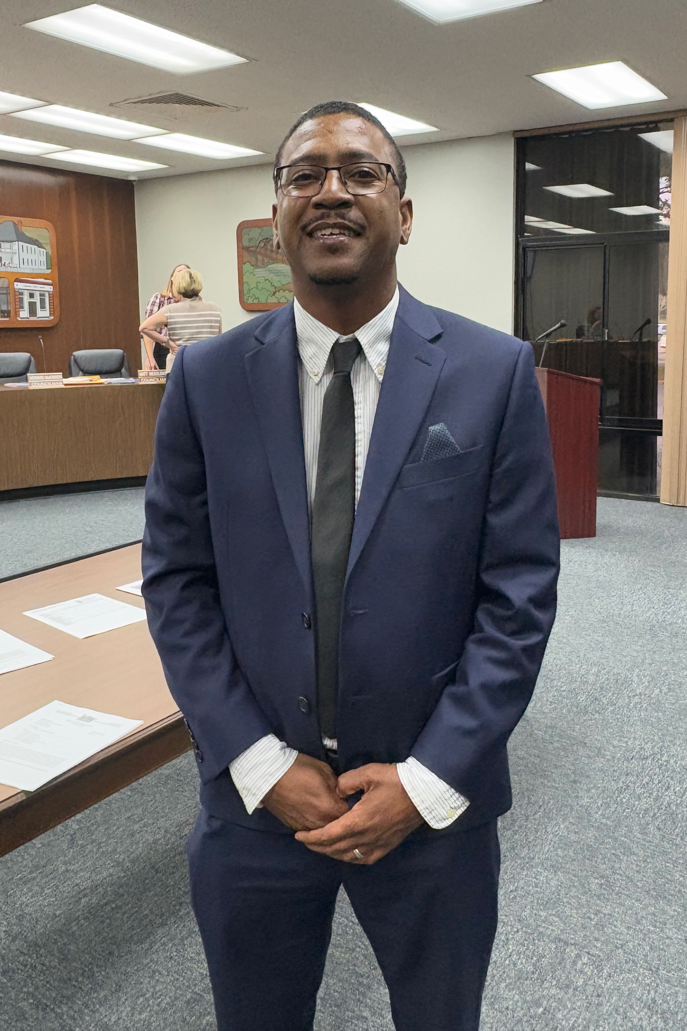 A man wearing a blue suit, white button down shirt, and charcoal-gray tie stands with a smile for a photo.