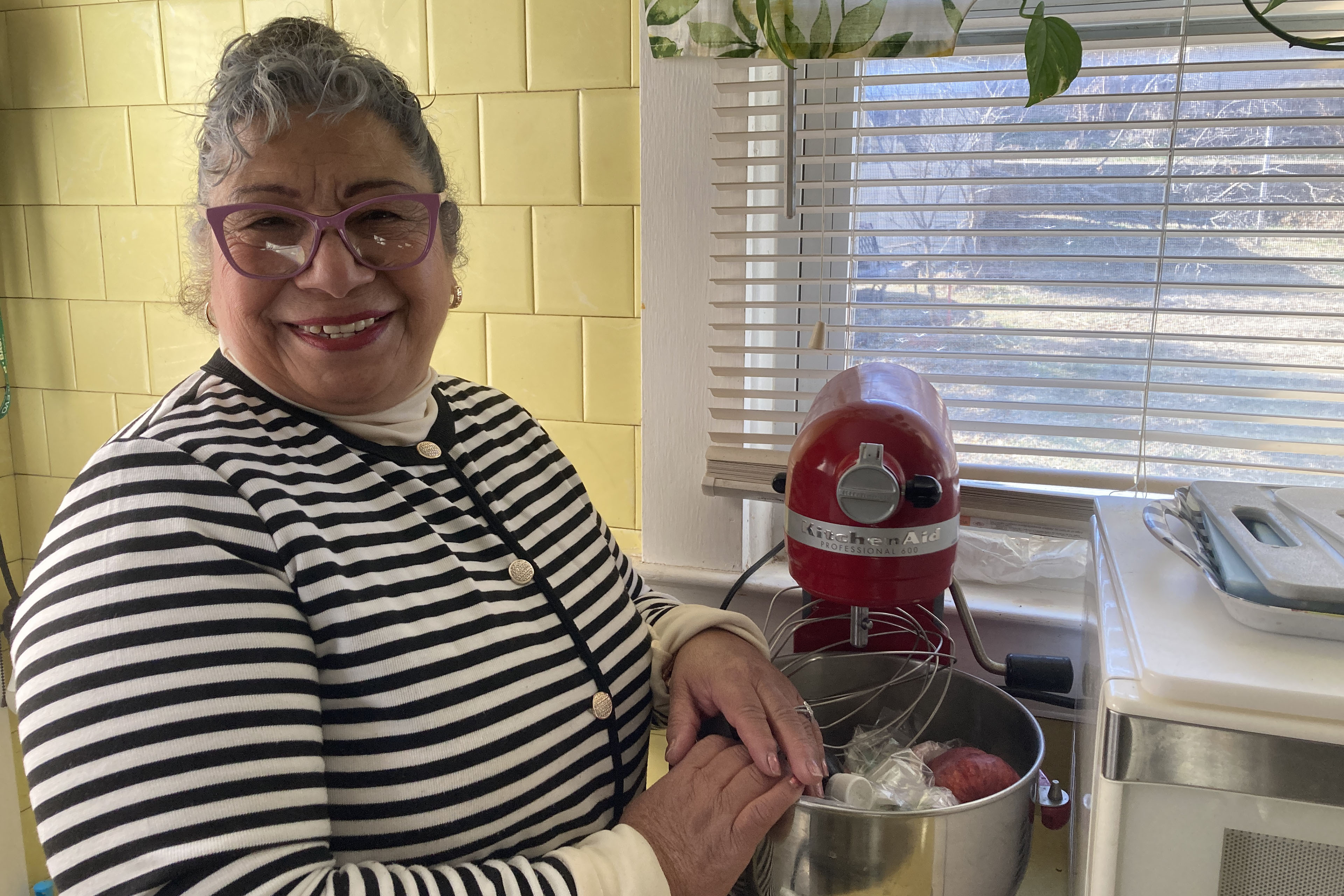 A woman wearing a black and white striped shirt and pink glasses smiles standing in a yellow-tiled kitchen