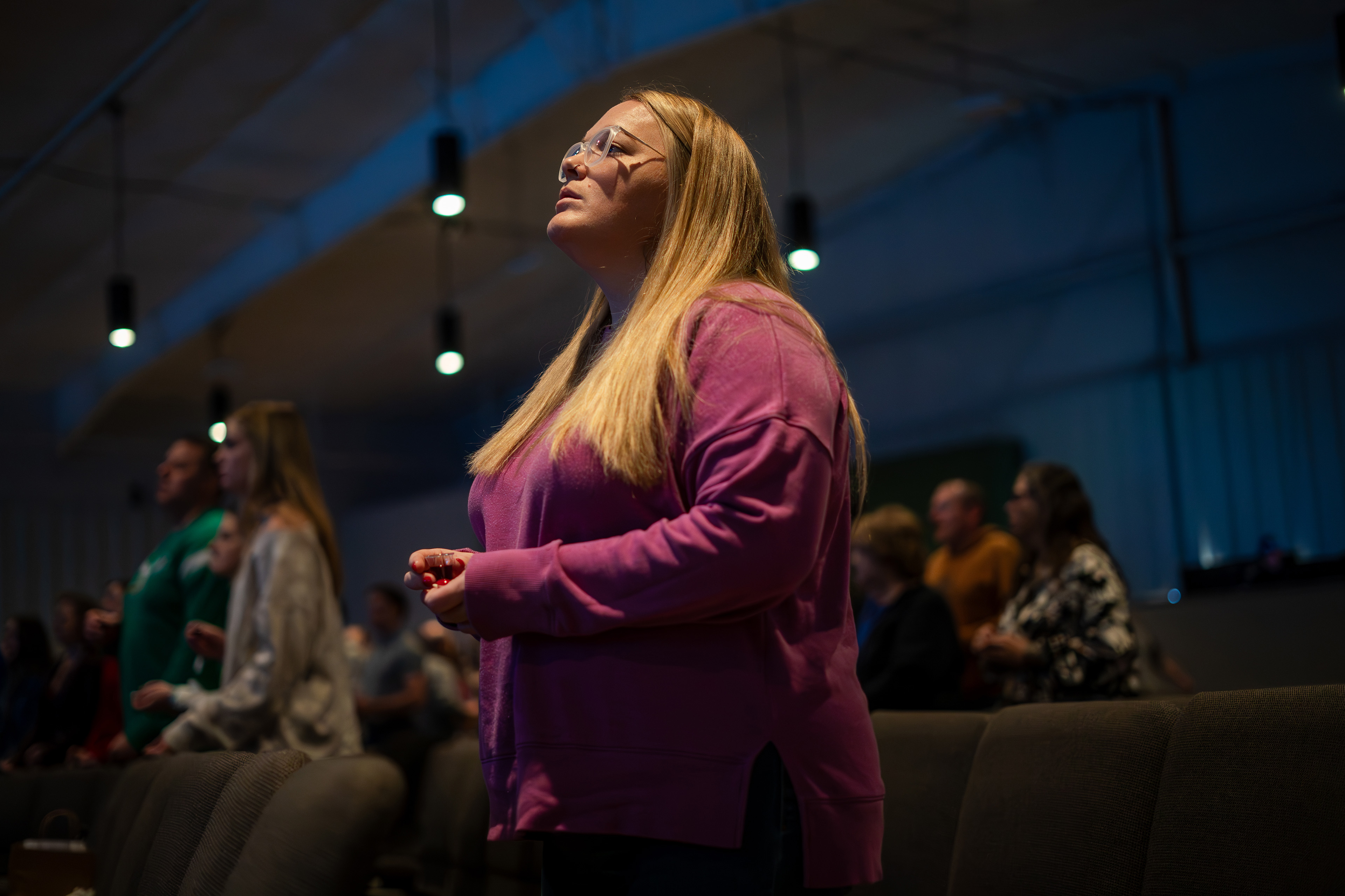 A blonde-haired woman wearing glasses stands in the pews at a church service