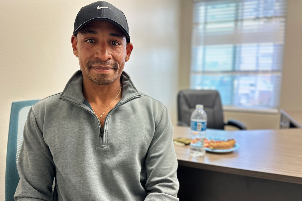 An adult man wearing a baseball cap and a high-collar sweatshirt looks into the camera.