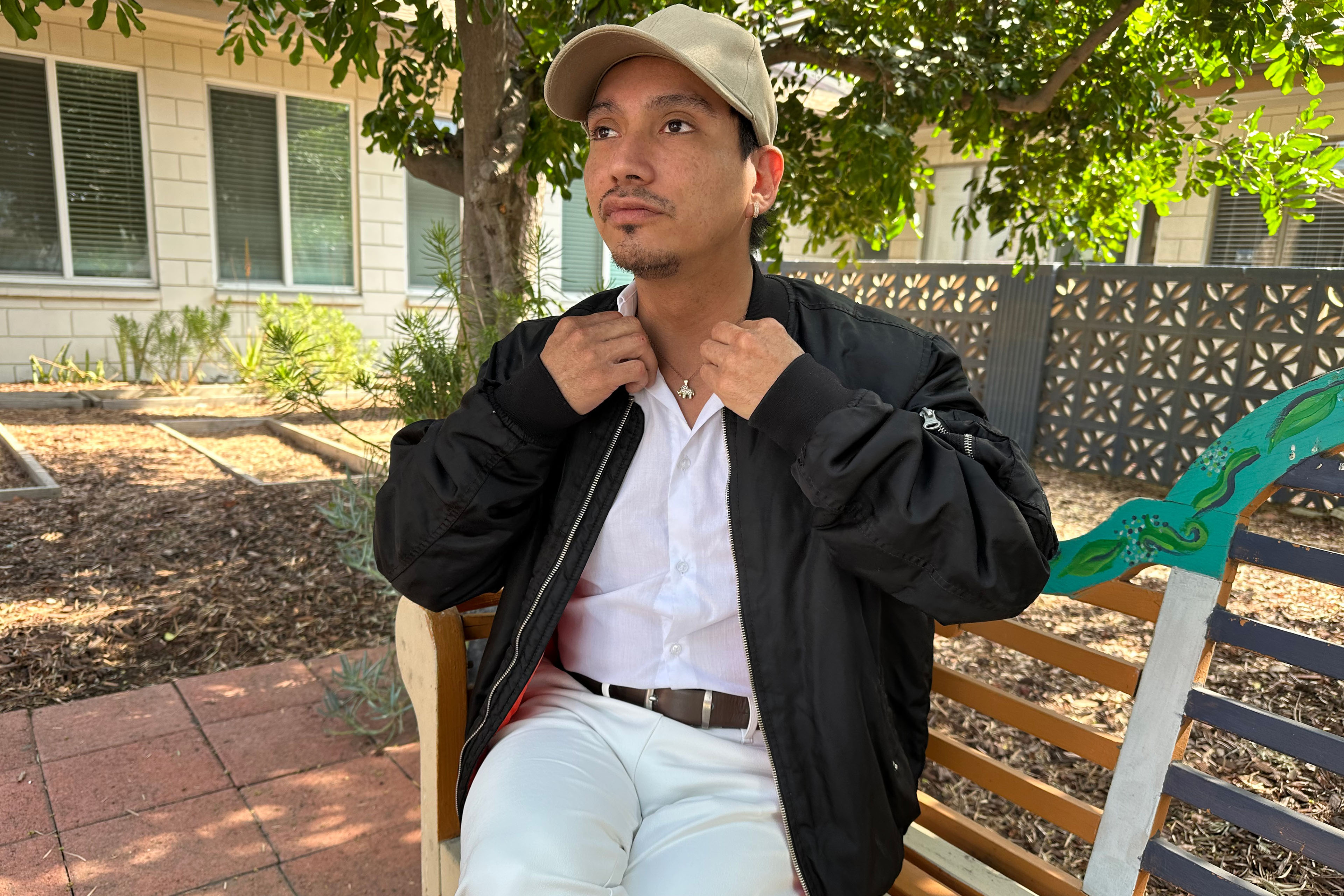 An adult man fixes his collar as he is photographed sitting on a bench outdoors on a sunny day.