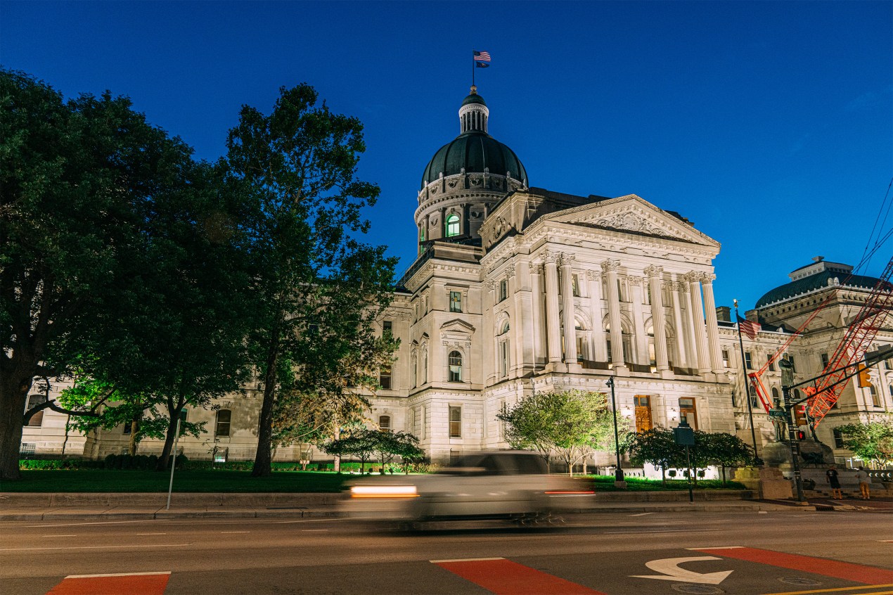 A photo of Indiana's Capitol taken at night as a car drives on the road in the foreground.