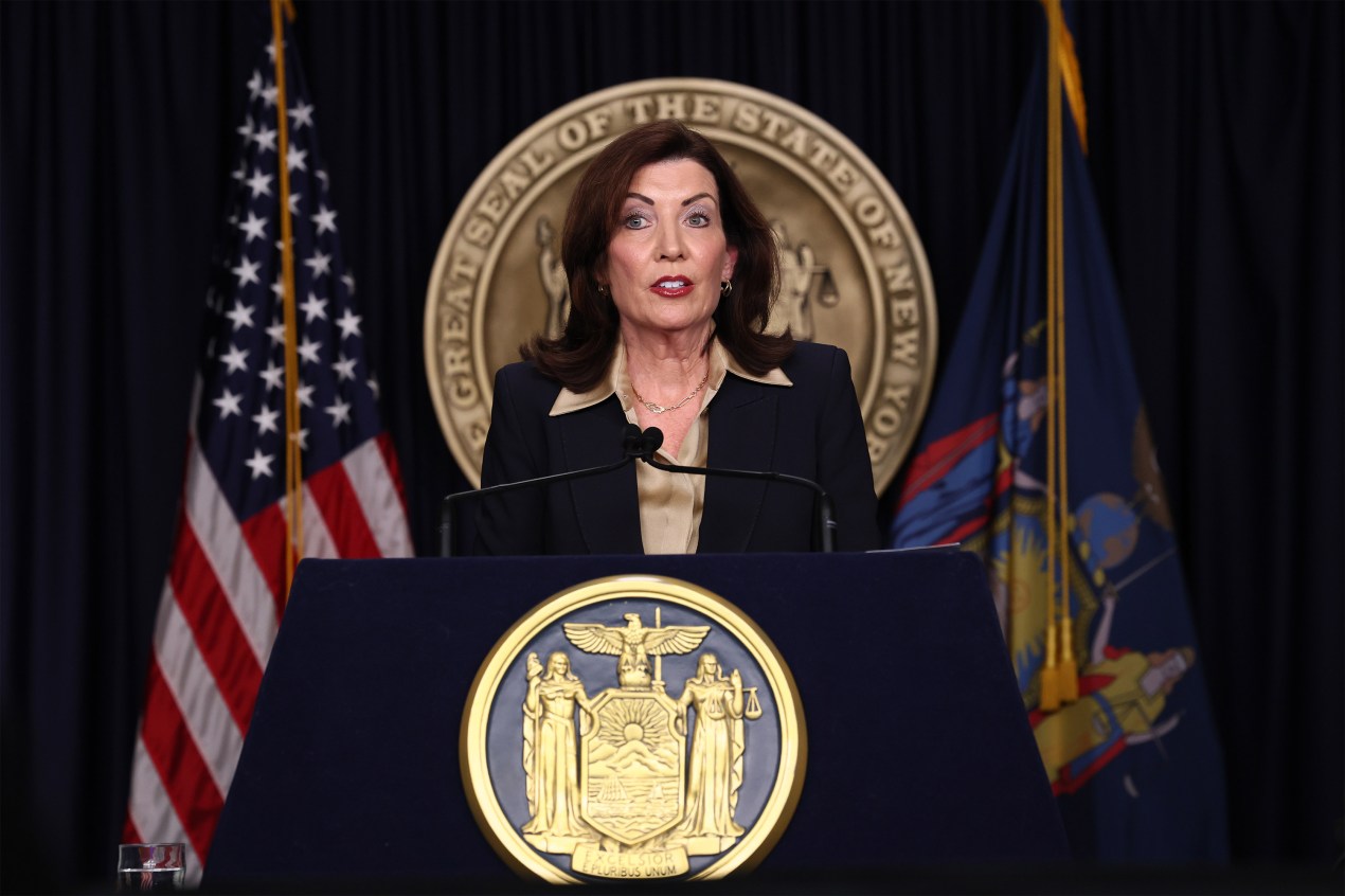 A photo of Kathy Hochul speaking at a podium with the USA and New York flags behind her.