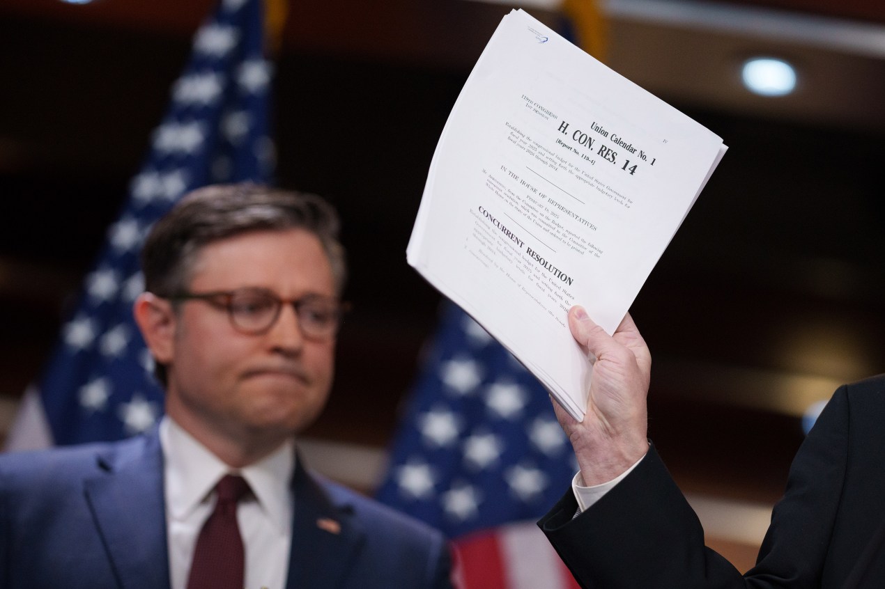 House Speaker Mike Johnson stands in the background of a press conference while a budget resolution bill is held up in front of him.