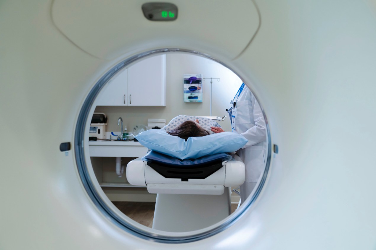 A photo taken from the inside of a CT scanner shows a patient about to be inserted into the device. A doctor stands at their side.