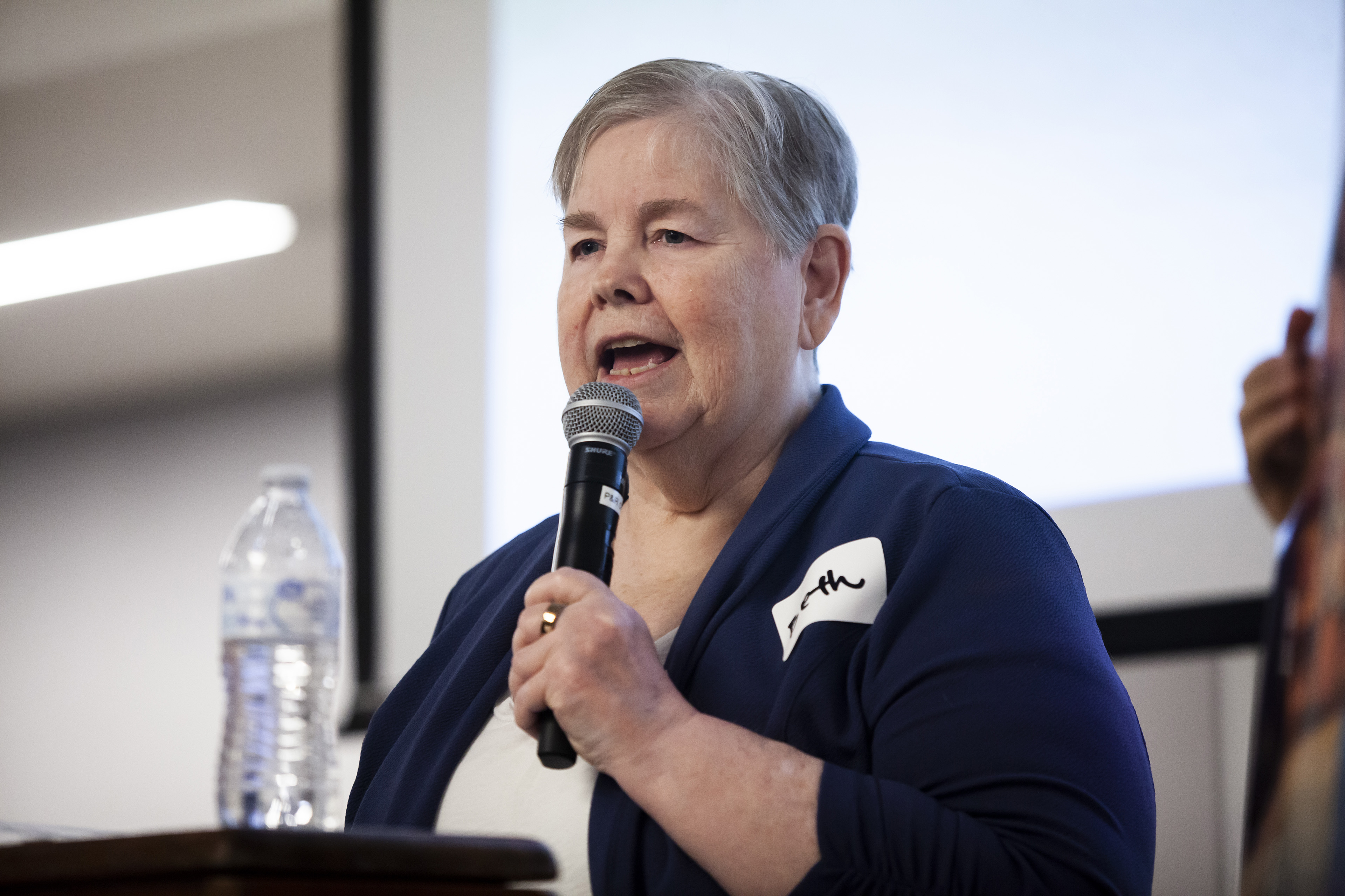 A woman with short and gray hair, wearing a navy sweater, speaks on a podium.