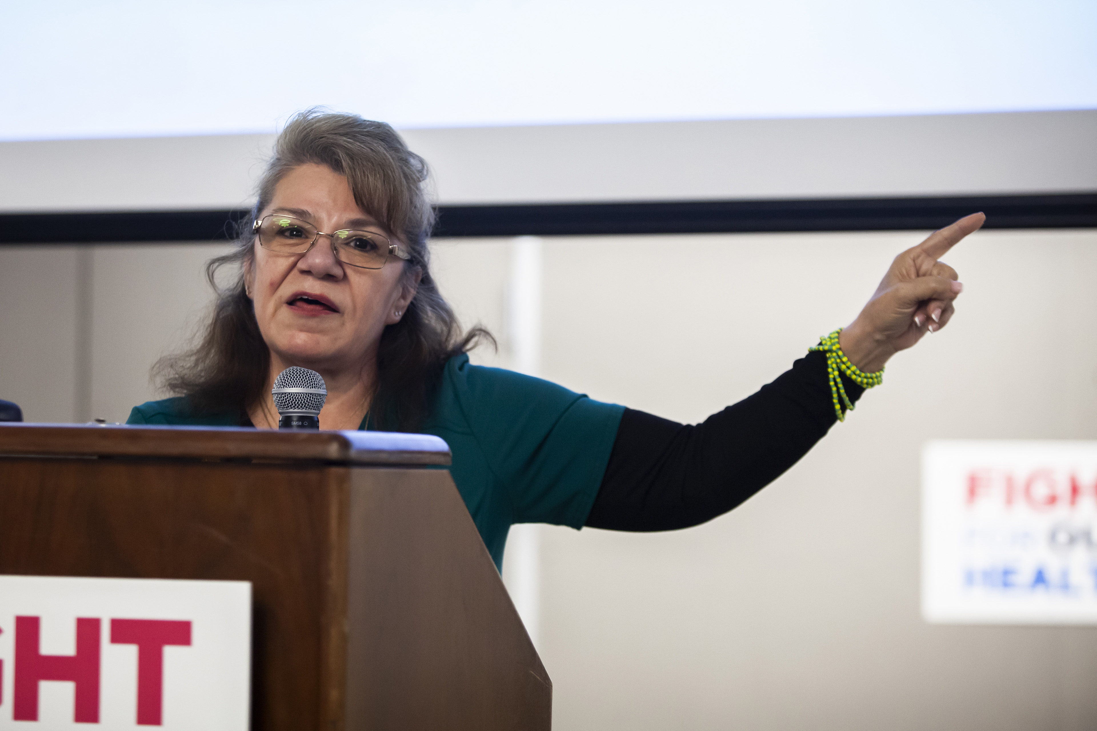 A woman wearing glasses stands at a podium and points her finger as she speaks.