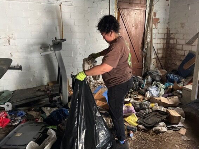 A photo of a woman filling a trash bag in a basement surrounded by clutter and debris.