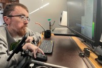 A photo of a man seated at a desk, working on a computer, breathing through a portable ventilator.