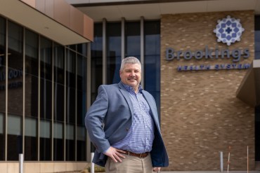 A man in business attire, with short-cut greying hair, stands in front of a hospital building. With his hands on his hips, he smiles. Behind him, building signage reads: "Brookings Health System"