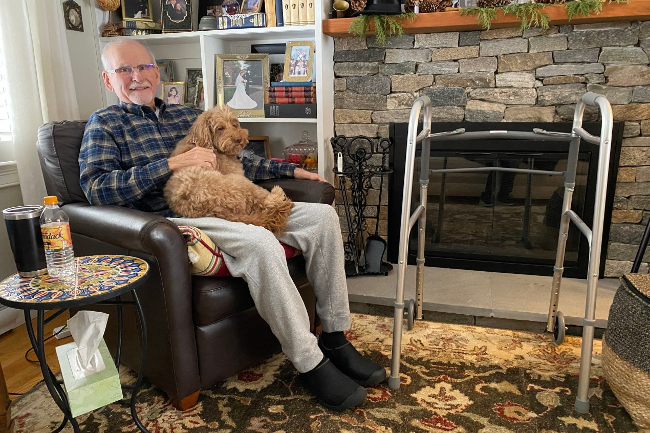 A senior man sits in a brown leather chair with a labradoodle dog in his lap. A walker is beside him. He is sitting in his home living room.