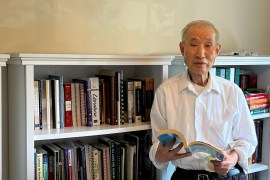 A senior man stands beside a bookshelf, holding a book. He wears a white button-shirt.