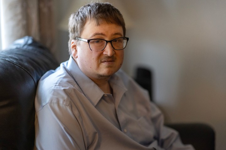 A man wearing glasses sits on a couch facing the camera.