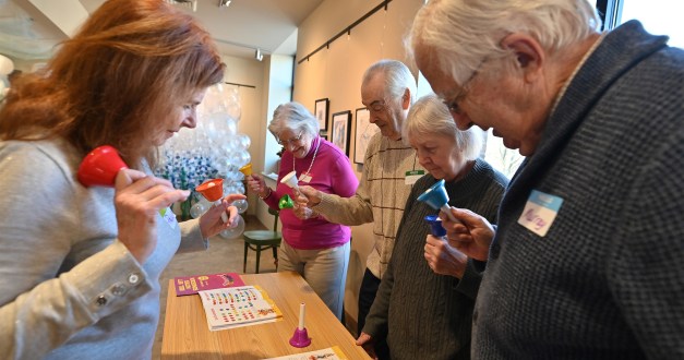 A photo of a woman leading a group of older adults in a game with hand bells.