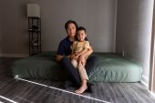 A young boy sits on his father's lap on a mattress covered in a green fitted sheet on the floor.