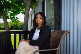 A woman sits on a wicker chair on an outdoor porch. She looks away from the camera, recalling a memory.