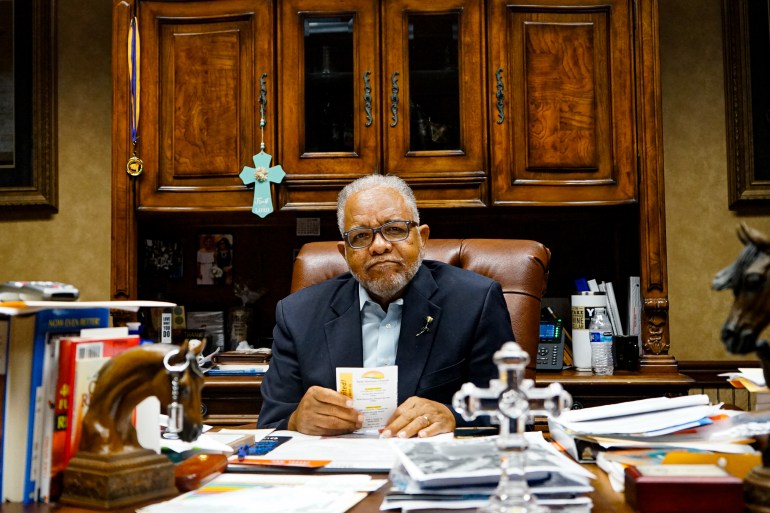 A man faces the camera as he sits his desk that is covered in papers and books.