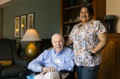 A senior man sits in a chair. A nurse stands beside him with her hand on his back, like good friends. They are both smiling in the direction of the camera.