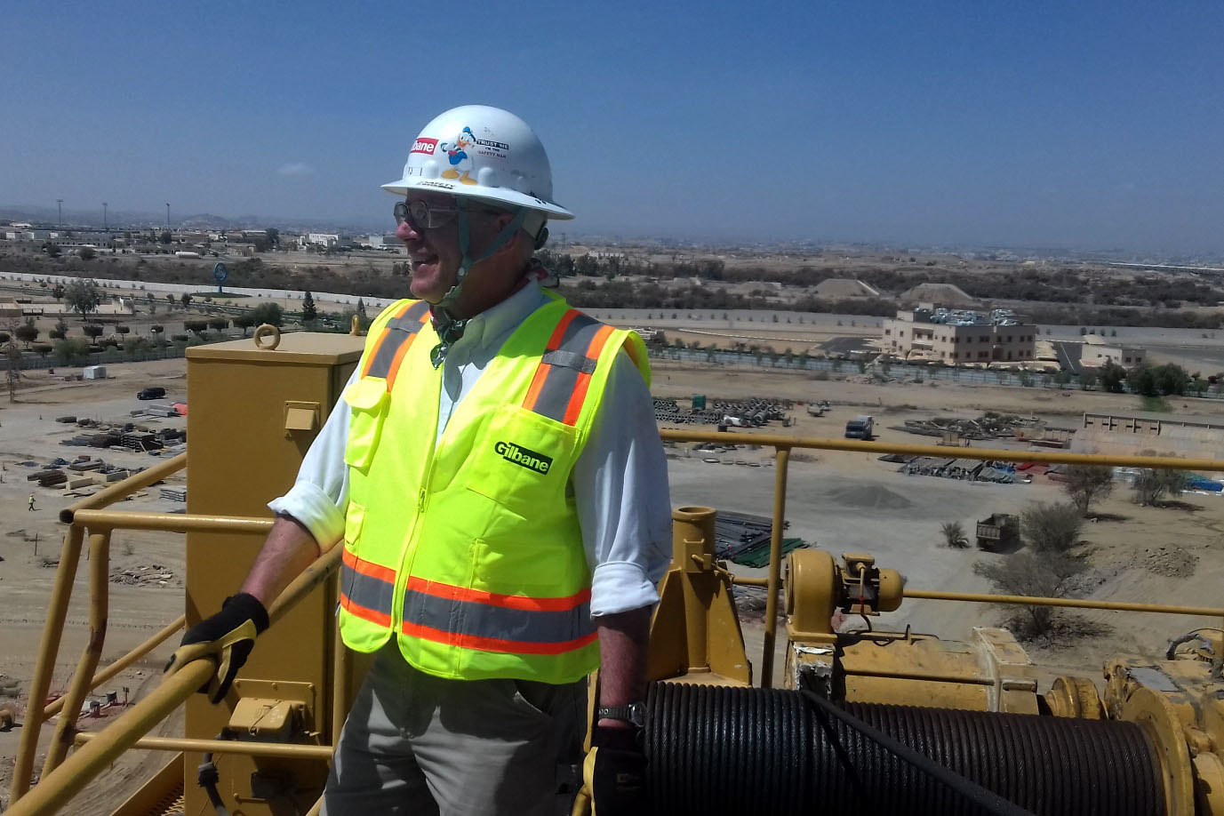 A photo of a man on a construction site. He stands on metal machinery that has been painted yellow, and wears a neon vest and a white hard-hat.