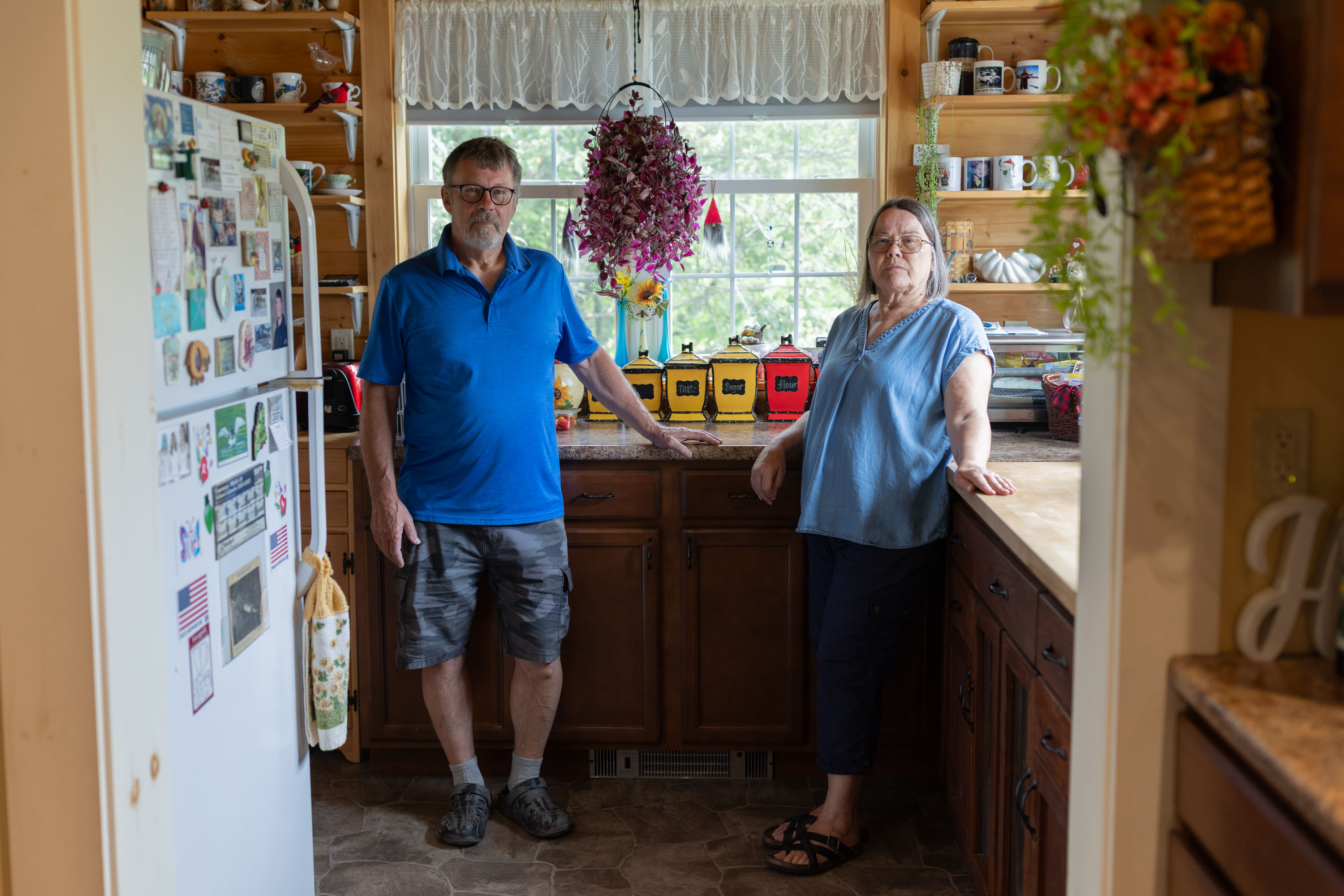 A husband and wife stand in their kitchen for a portrait. The kitchen has a familial warmth to it; flowers hang from baskets, the fridge is covered in photos and magnets, and colorful jars and mugs line the shelves.
