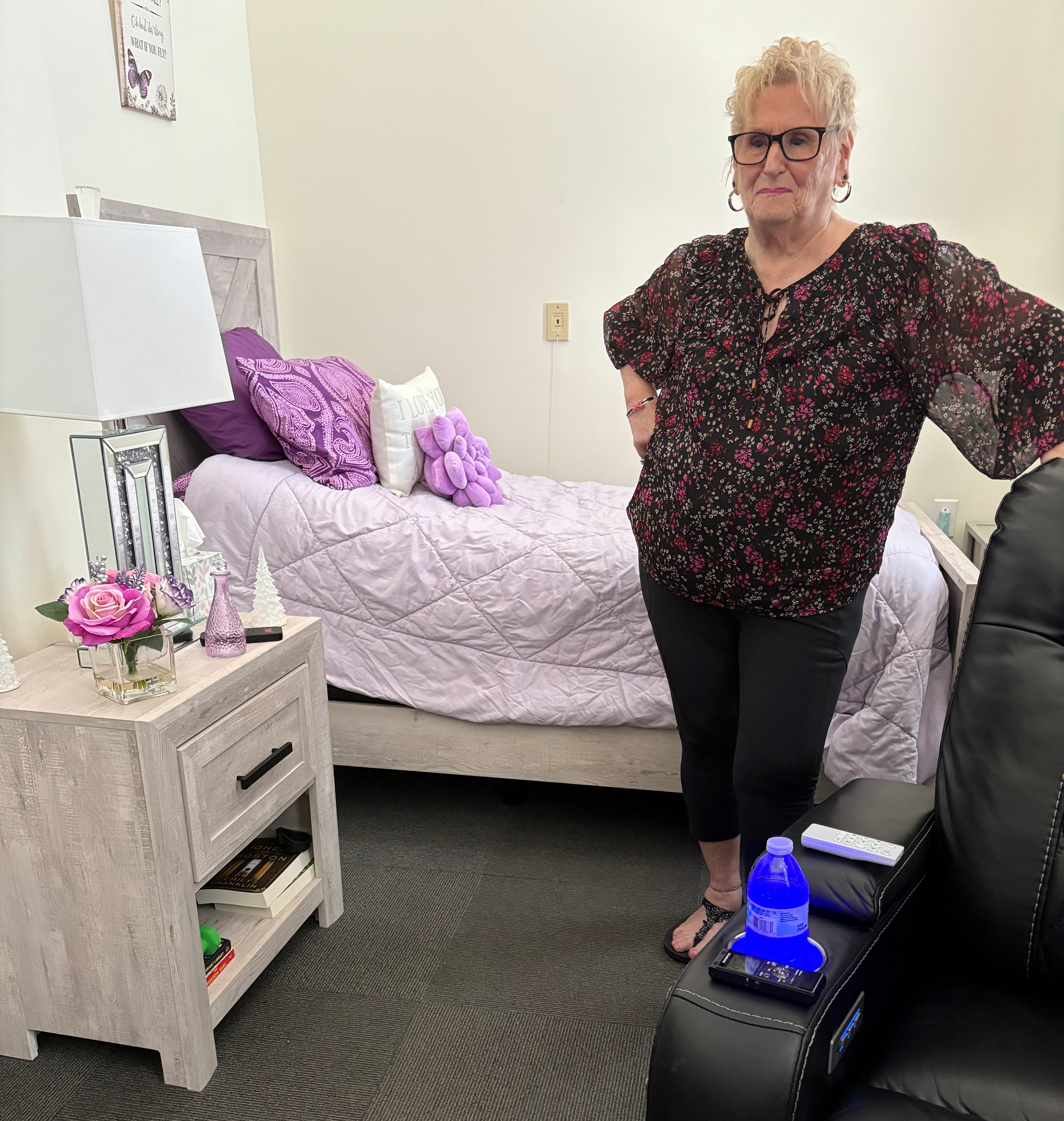 Ein Foto von Roberta Rabinovitz in ihrem Studio-Apartment. Die Tagesdecke und die Kissen hinter ihr sind lila.