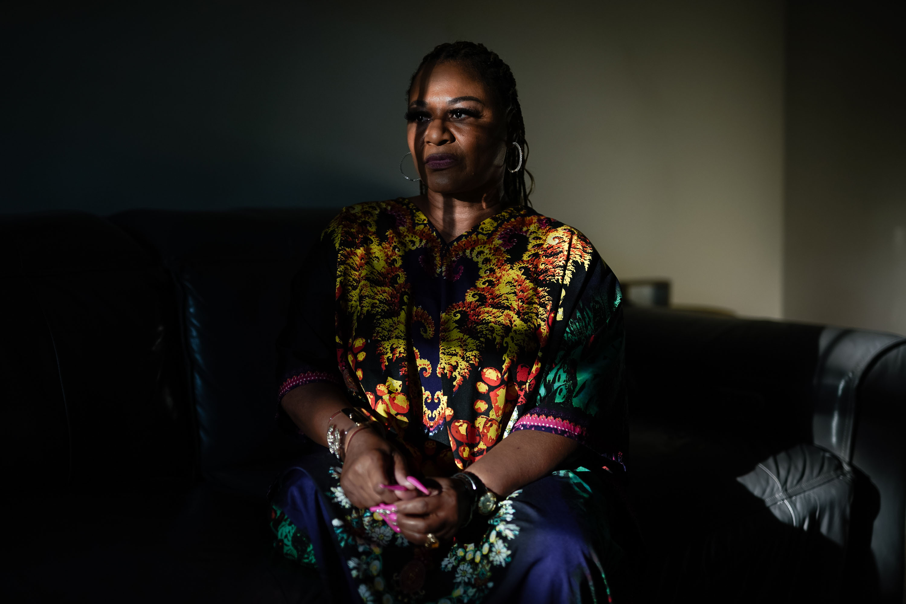A portrait of a woman sitting on a couch in a dimly lit room. A shadow from a window pane crosses over her face dramatically.
