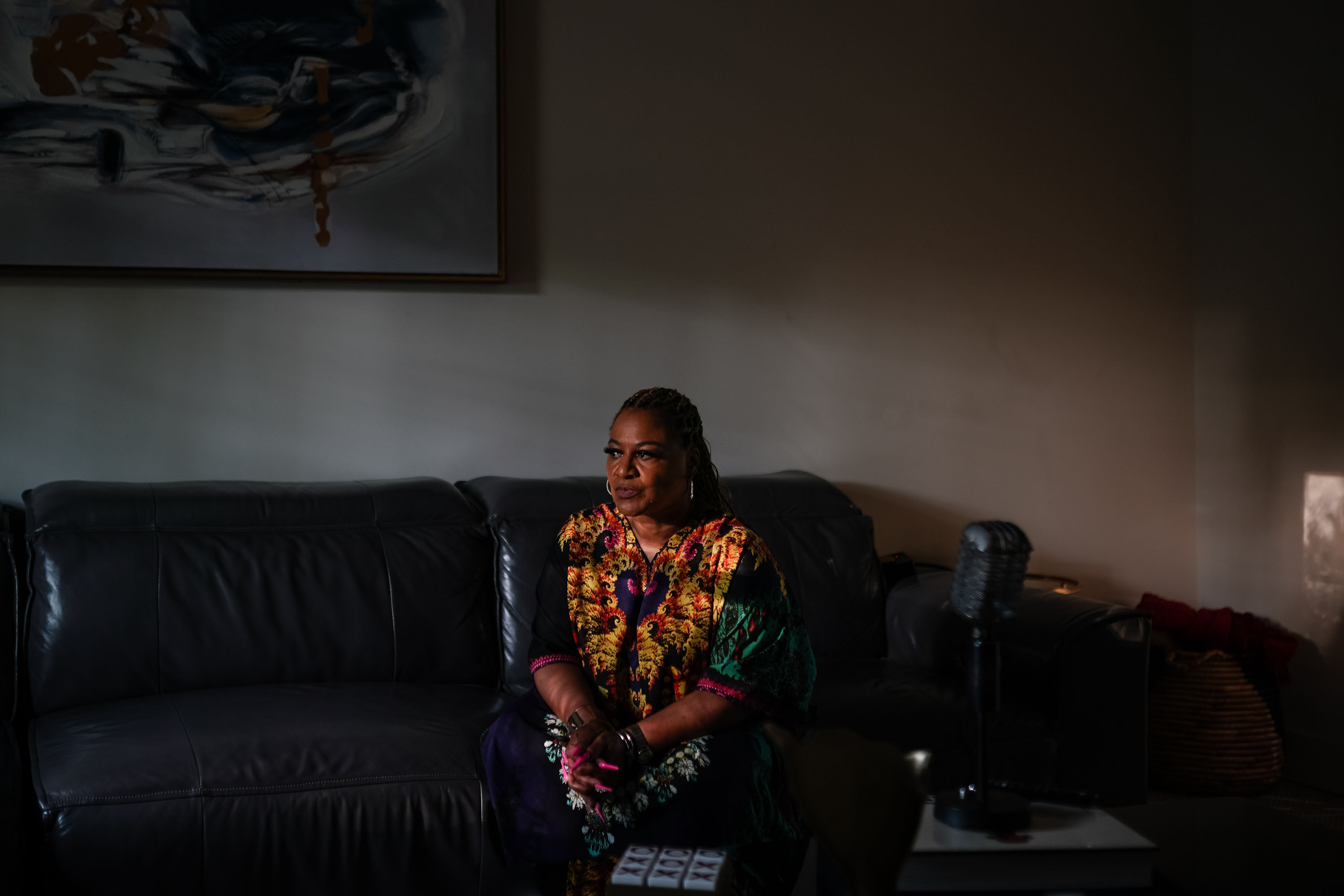 A portrait of a woman sitting on a couch in a dimly lit room. She holds her hands together in her lap and has a serious expression.