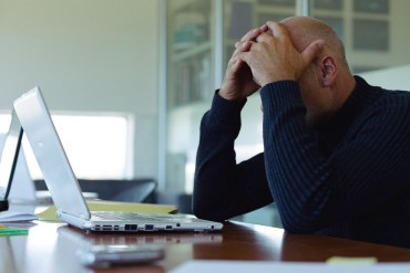 A photo of man holding his head in his hands as he uses a laptop.