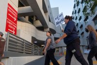 Four people are in frame walking past a sign with white lettering on a red background reading "Emergency Entrance" and "Emergency Department Chest Pain Center" each with arrows pointing right. One person carries a handwritten sign that reads "Keep ICE Out of Hospitals."
