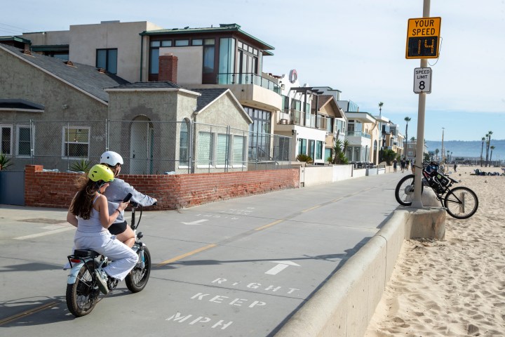 A photo shows a pair riding an e-bike across The Strand, a paved road in Hermosa Beach, California. The speed limit is posted at 8 miles per hour, and a speedometer shows they are going 14 miles per hour.
