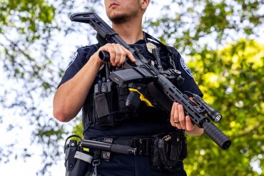 A police officer holding a rifle stands outside.