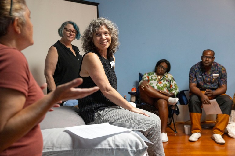 A woman sits on a table during an aid demonstration. She smiles as she looks at another woman who is speaking.