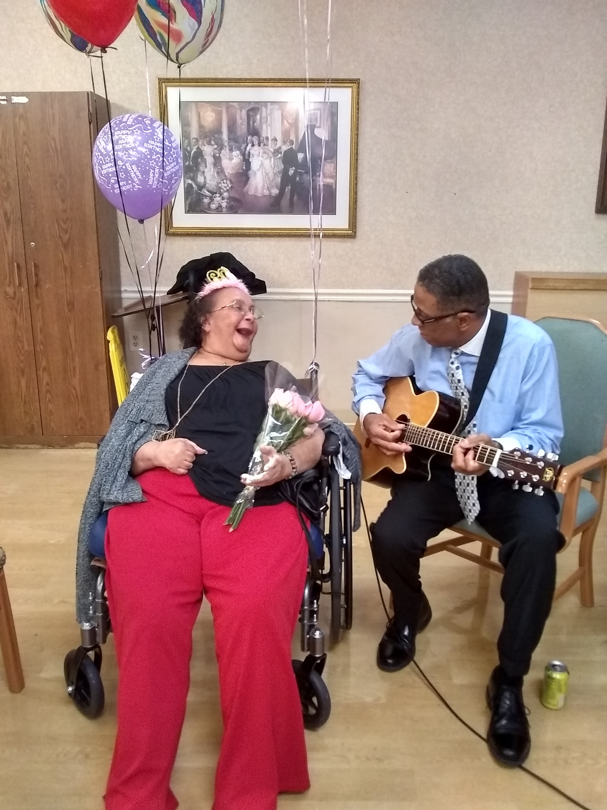 A woman wearing a crown and holding flowers watches a man play guitar next to her