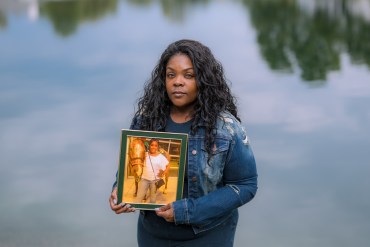 A photo of a Black woman standing by water. She holds a framed photo of her deceased mother.