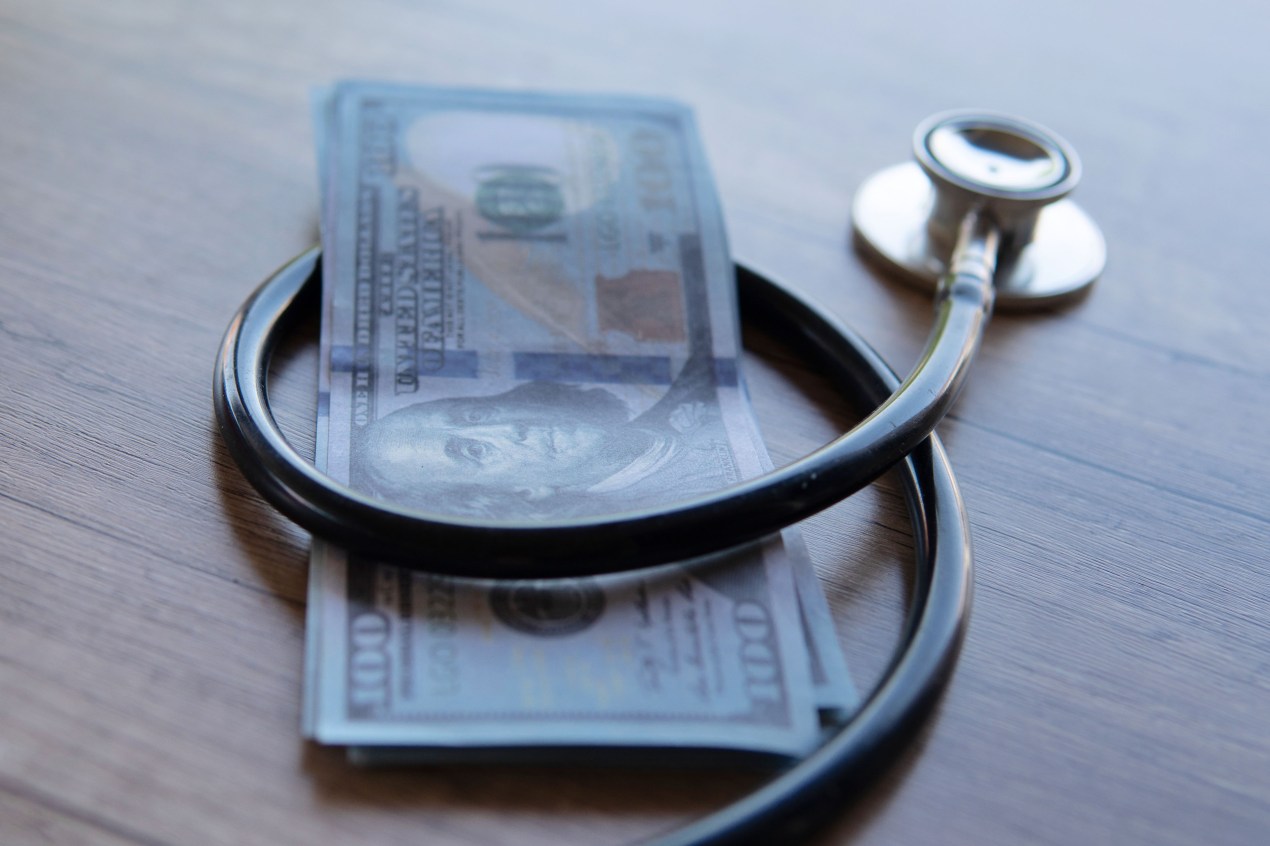 A close-up image of a black stethoscope wrapped around a stack of U.S. dollar bills.