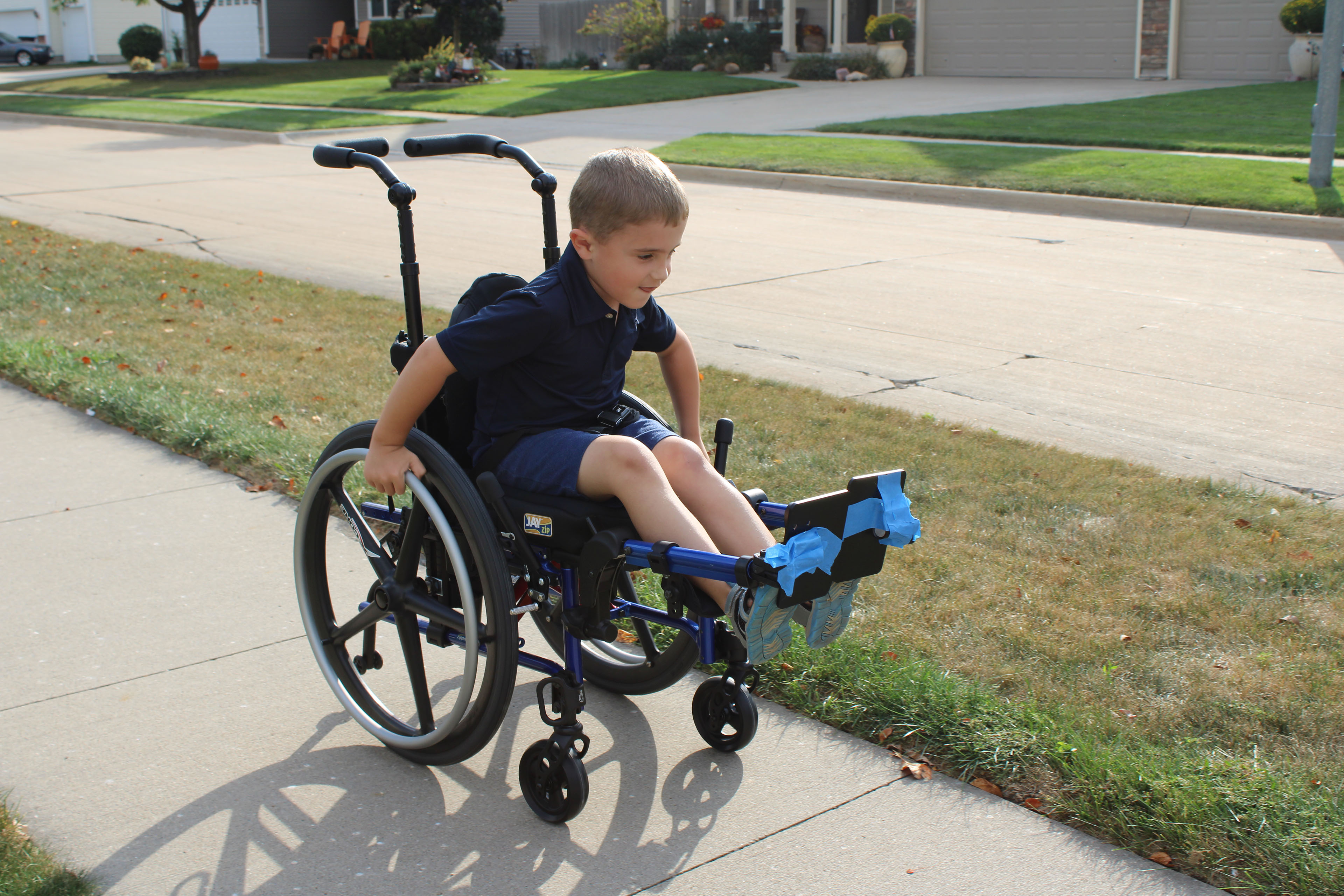 A young boy in a wheelchair smiles as he rolls himself down a sidewalk on a sunny day.