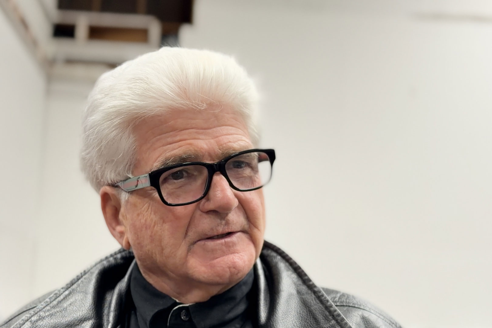 A man with white hair and dark glasses speaks to someone just off-camera.