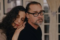 A woman with dark hair leans with her eyes closed on the back of a man wearing glasses who looks at the camera