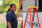 A man with long dark hair pulled into a ponytail and wearing a dark blue t-shirt stands near a building under construction.